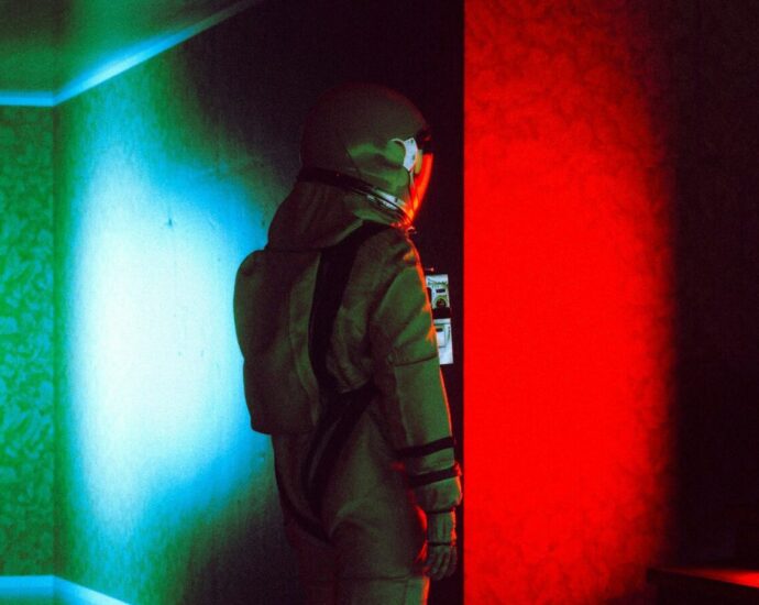 Person wearing a space suit standing in a dark room with the colors red and green reflecting on the walls.